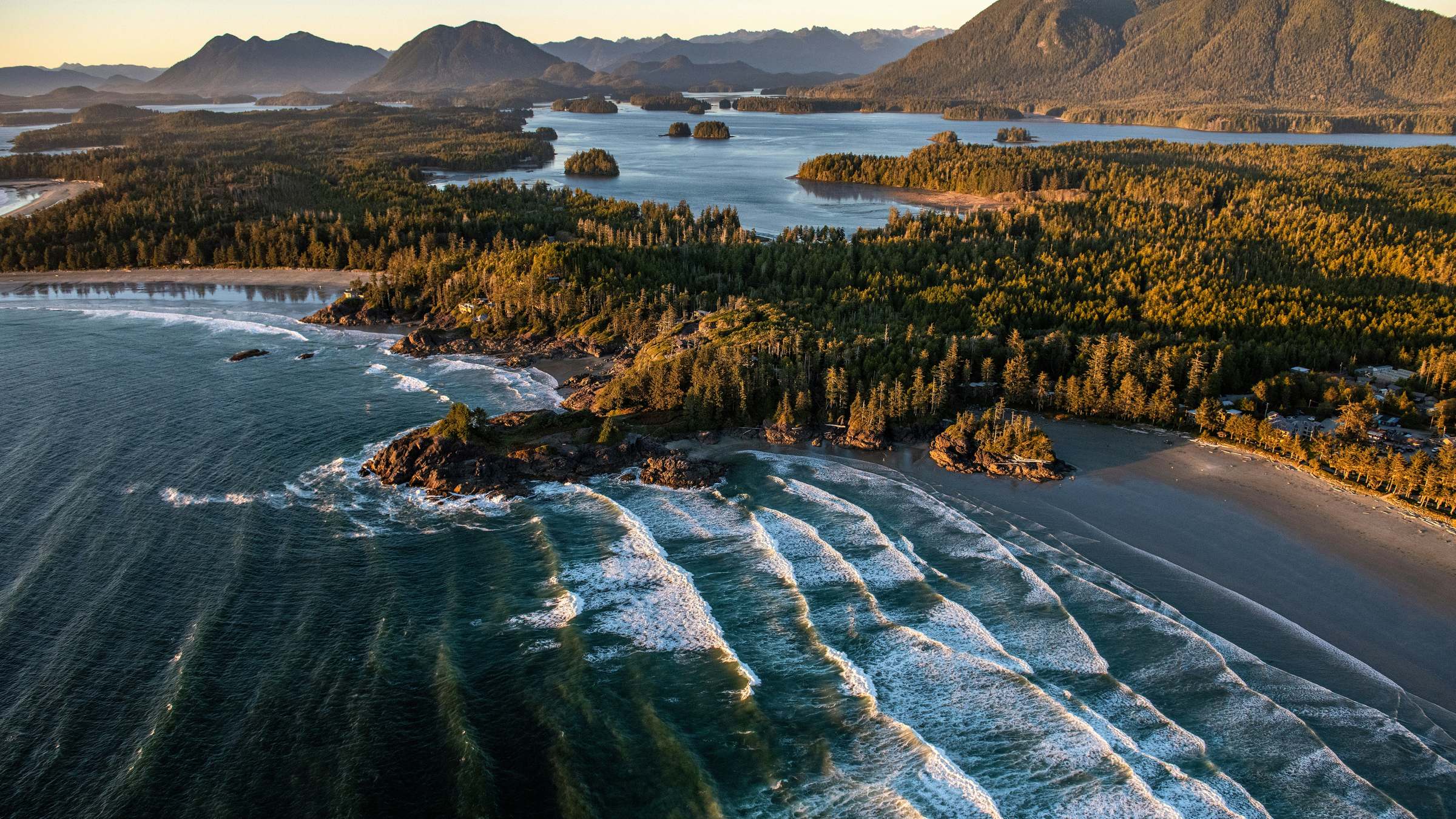 Aerial view of a Pacific coastline at golden hour: forested islands, breaking waves, distant mountains.